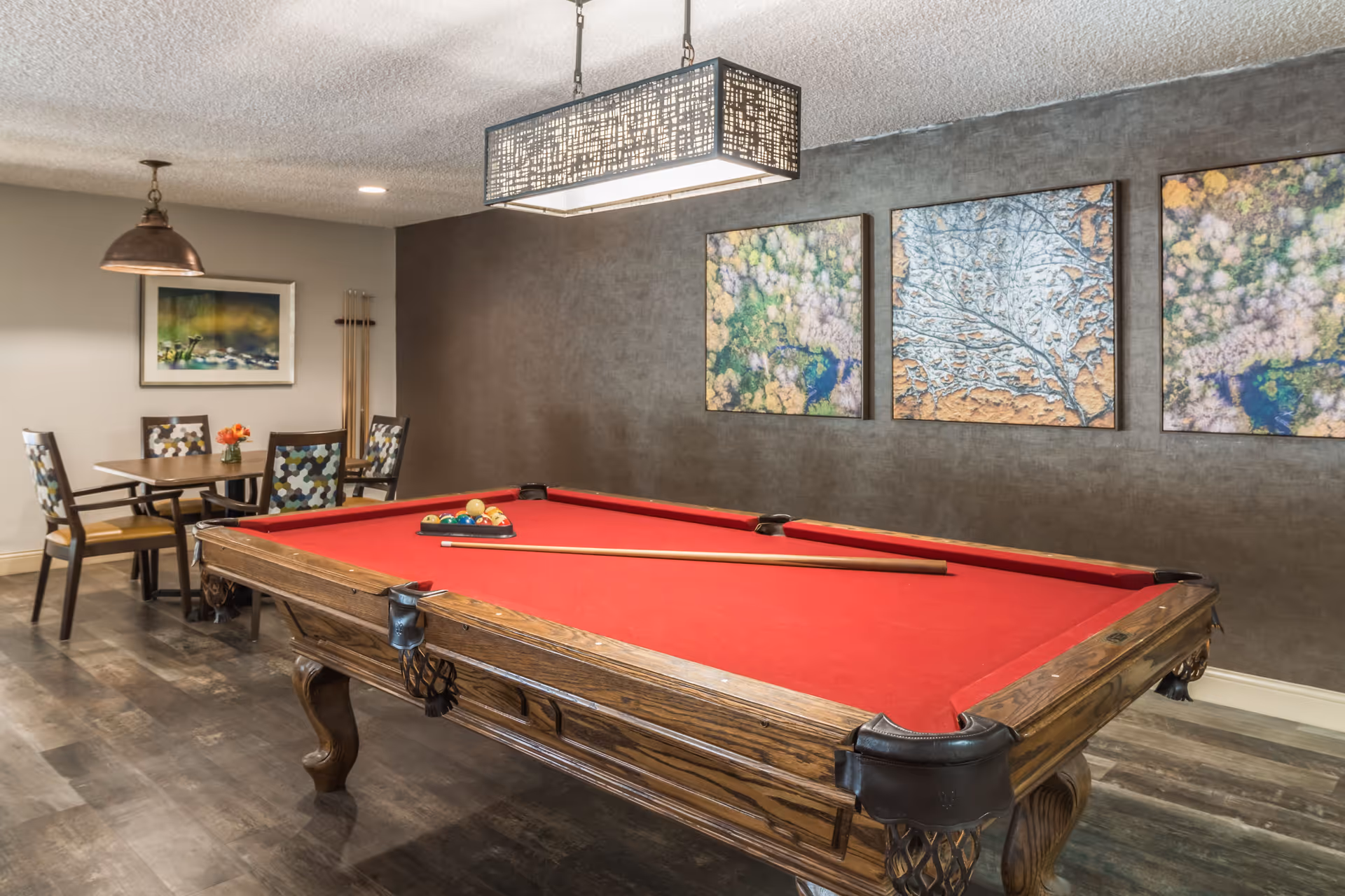 A recreational room featuring a wooden pool table with a red felt surface, a cue stick resting on it, and a rack of billiard balls. Behind the pool table, there is a small dining area with a table and four chairs with patterned upholstery. The walls are decorated with three abstract paintings, and the room has modern lighting fixtures hanging from the ceiling.