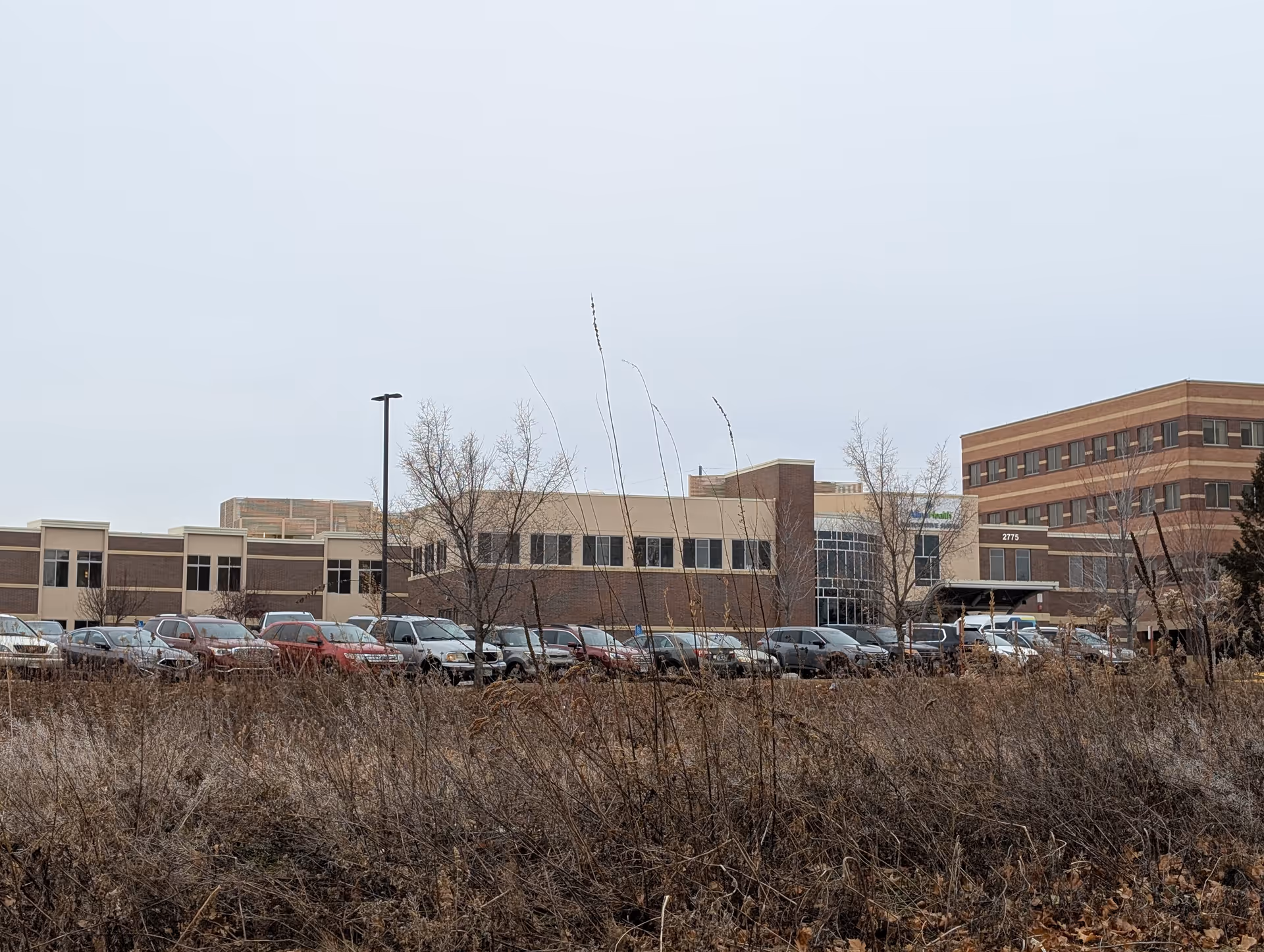 Exterior view of a large multi-story building with a parking lot full of cars in front. The building has a combination of beige and brown brick walls with many windows. Leafless trees and dry grass are visible in the foreground under an overcast sky.