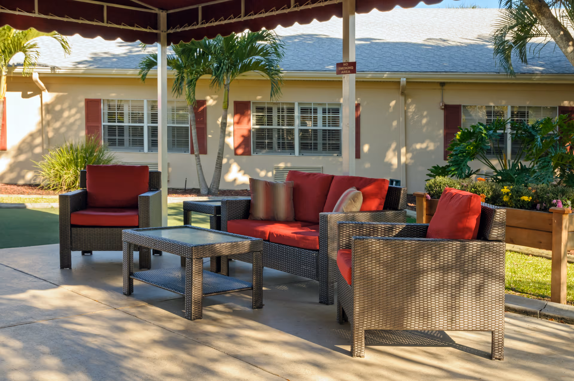 Outdoor seating area with wicker furniture including a sofa and two armchairs with red cushions, and a glass-top coffee table under a shaded canopy. The background shows a building with windows, red shutters, palm trees, and landscaped greenery.