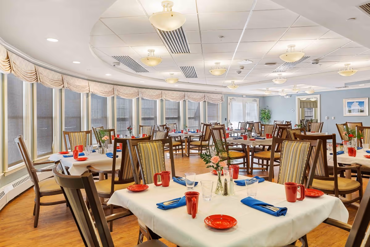 Bright dining room with multiple tables set with white tablecloths, red mugs, blue napkins, and wooden chairs under ceiling lights and large windows.
