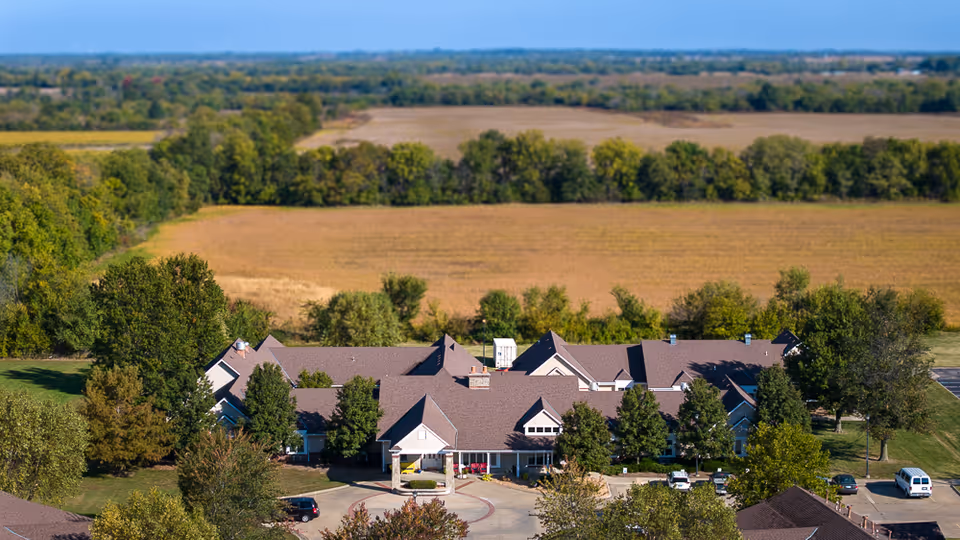 Aerial view of a single-story senior living facility with a circular driveway surrounded by trees and open farmland.