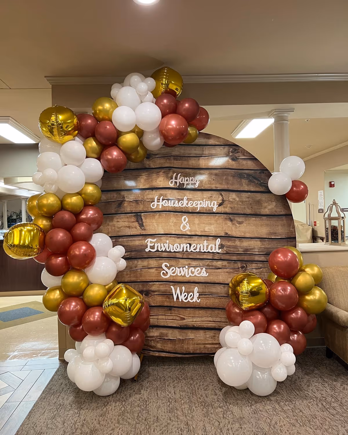A large circular wooden backdrop decorated with clusters of white, gold, and maroon balloons. The backdrop has white text that reads 'Happy Housekeeping & Environmental Services Week'. The setting appears to be an indoor common area with carpeted flooring and beige walls.