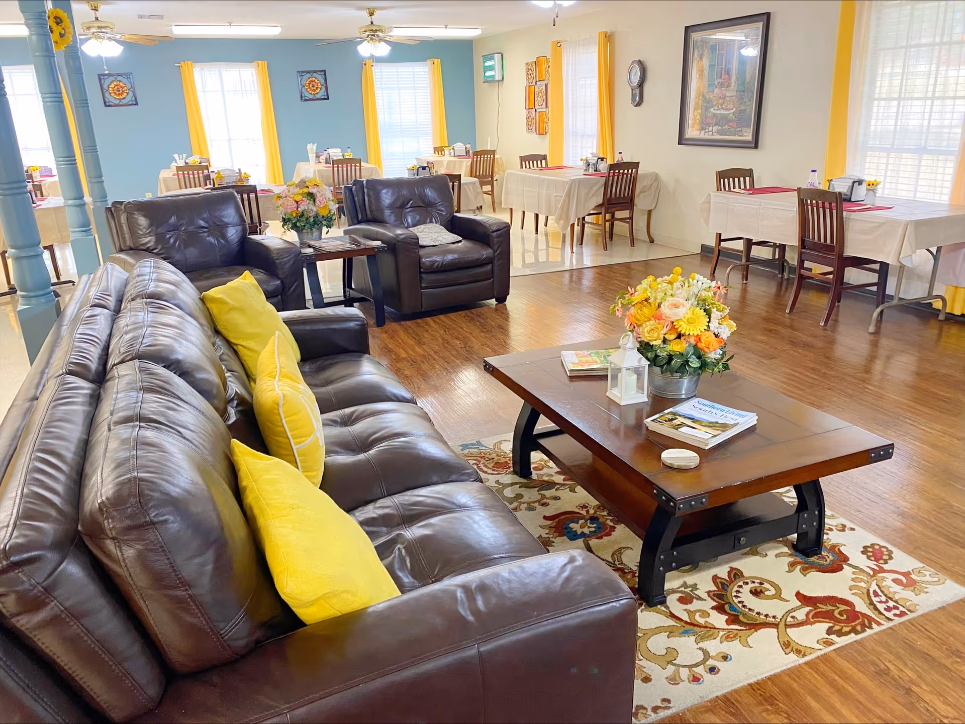 A bright and cozy common area in an assisted living facility featuring a dark brown leather sofa with yellow pillows, two matching armchairs, and a wooden coffee table with a floral arrangement and books. In the background, there are several dining tables with chairs, set against walls with yellow curtains and blue and cream paint. The floor is a mix of wood and tile, and the room is well-lit with ceiling fans and natural light from windows.