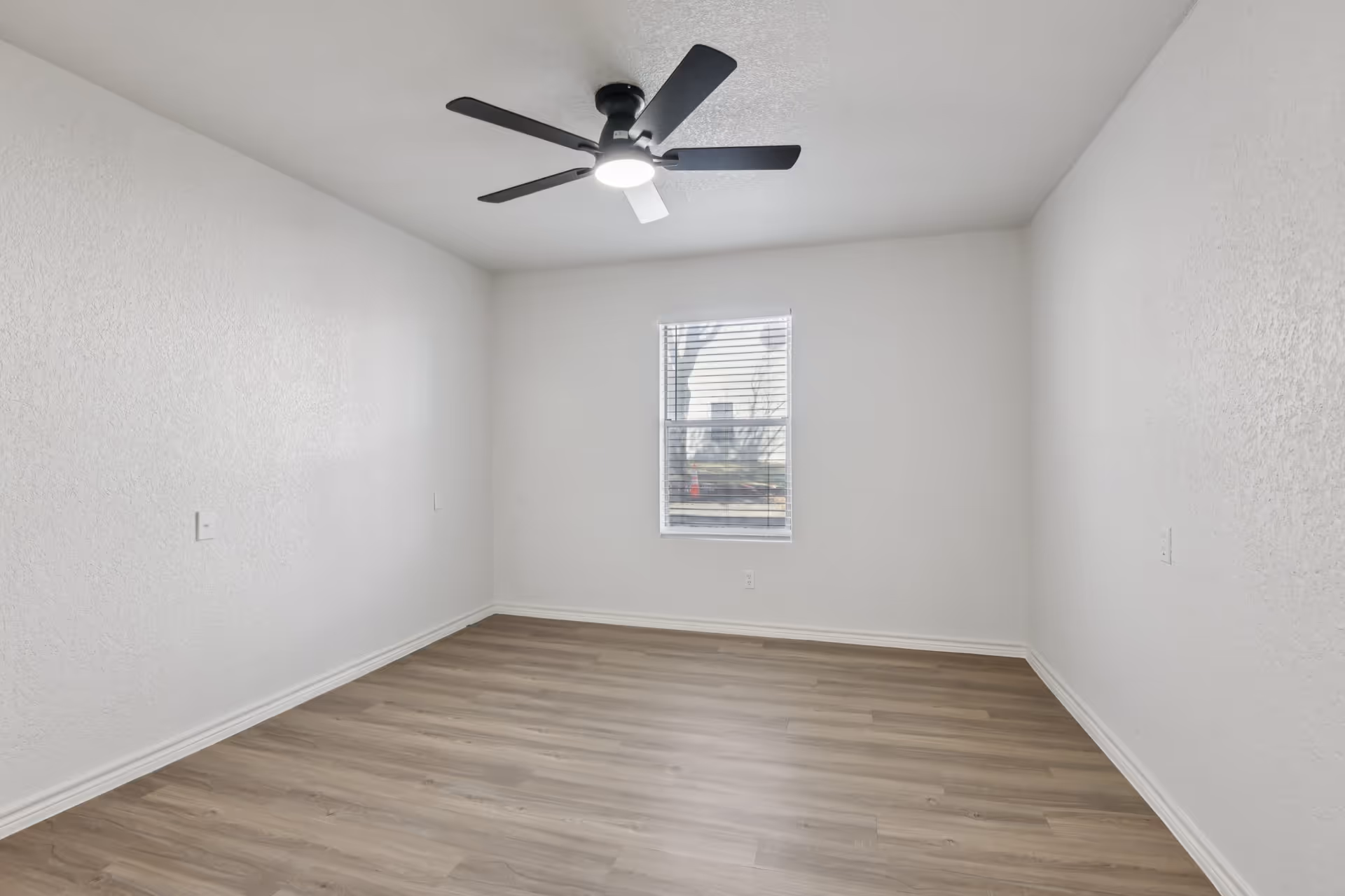 Empty white-walled room with a ceiling fan, small window, and light wood flooring.