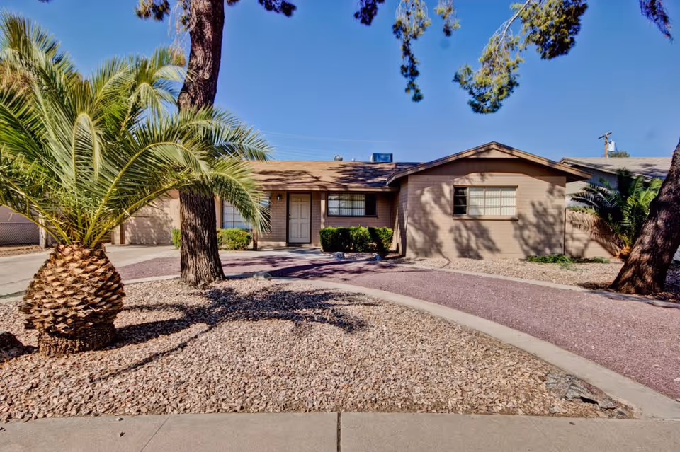 Single-story beige house with desert landscaping, palm tree, and a curved gravel driveway under a clear blue sky.