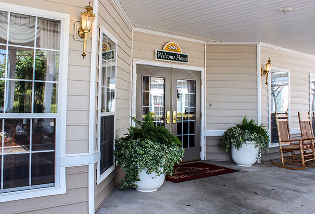 Entrance to a building with double glass doors, a 'Welcome Home' sign above the doors, two large white planters with green plants on either side of the entrance, beige siding, two wall-mounted lantern lights, and wooden rocking chairs to the right.