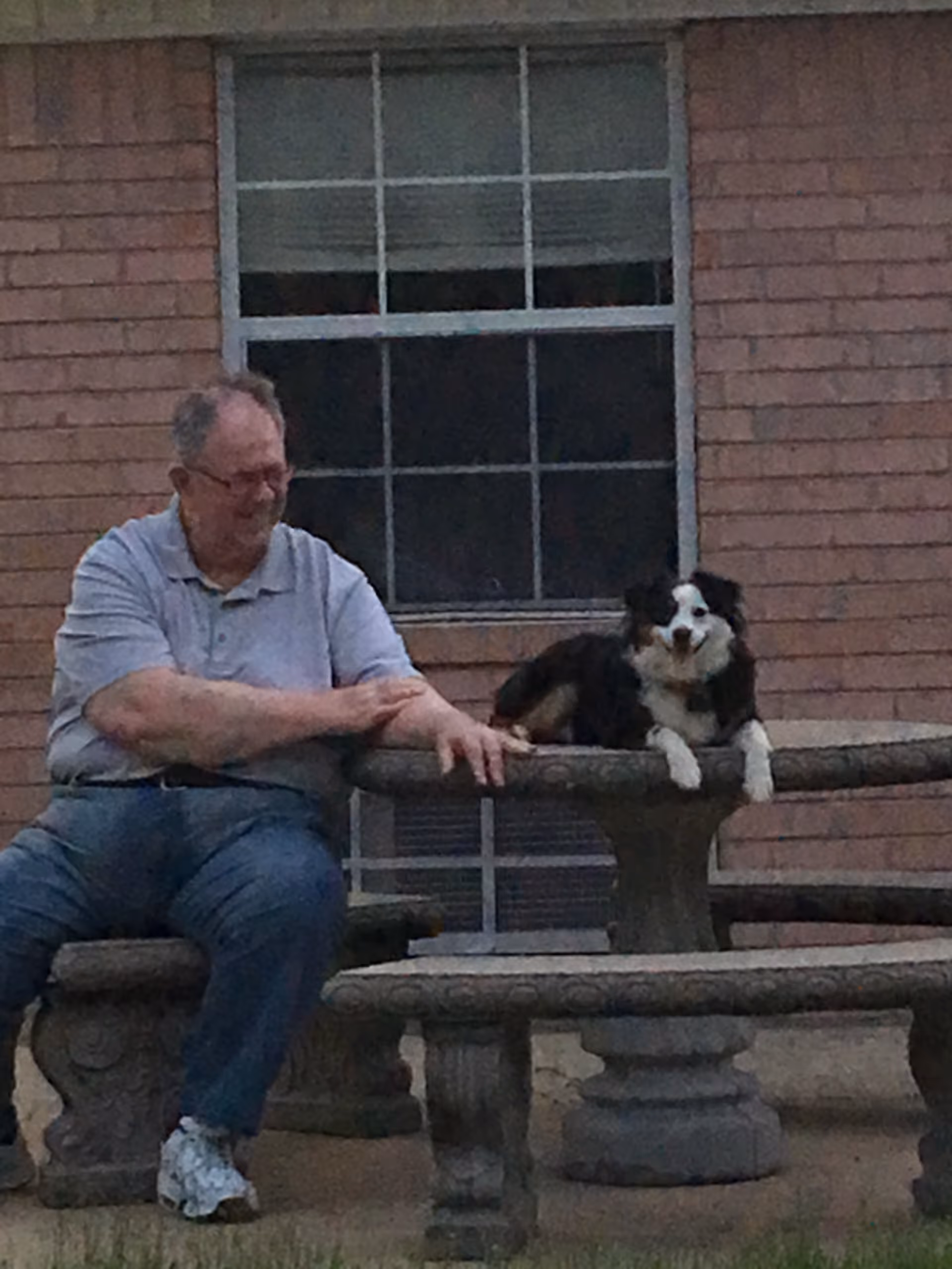 A man sits on a stone bench while a dog rests on a round stone table outside a brick building with a window.