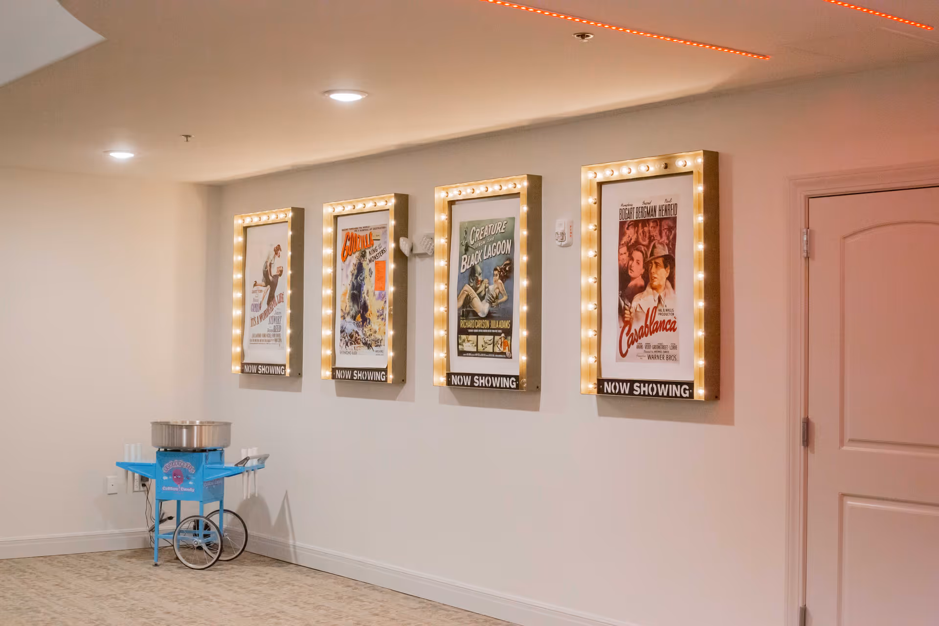 A hallway with four illuminated vintage movie posters on the wall, each framed with light bulbs and labeled 'NOW SHOWING.' Below the posters is a blue cotton candy machine on wheels with cups placed on its side. The floor is carpeted, and there is a closed door to the right.