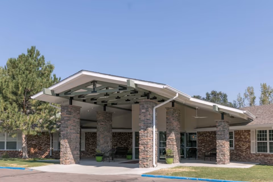 Entrance of a single-story brick building with a covered driveway supported by stone pillars, surrounded by green grass and trees under a clear blue sky.