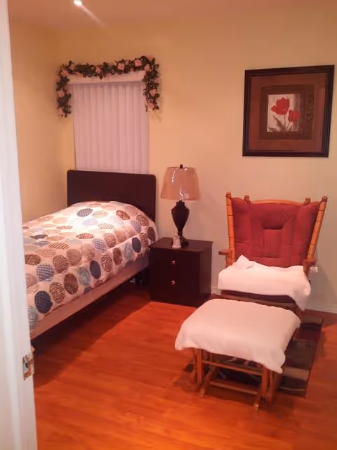 A cozy bedroom with a single bed covered in a quilt with circular patterns, a dark wooden nightstand with a lamp, a cushioned wooden rocking chair with a matching footrest, and a framed floral picture on the wall. The room has wooden flooring and light-colored walls.