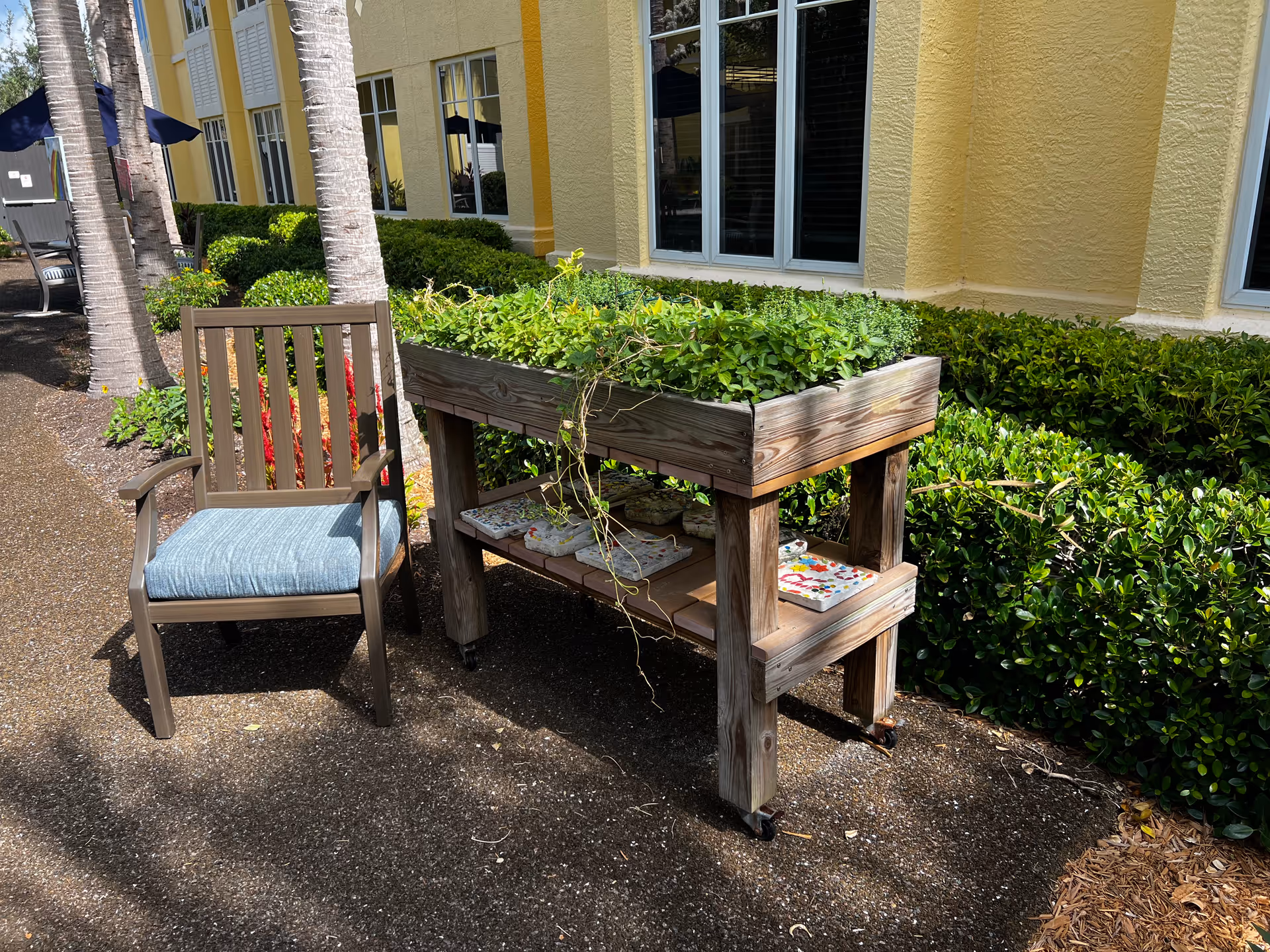 Outdoor scene showing a wooden chair with a blue cushion next to a raised wooden planter filled with green plants. The planter has a lower shelf with decorative tiles. The background features a yellow building wall with windows and neatly trimmed bushes.