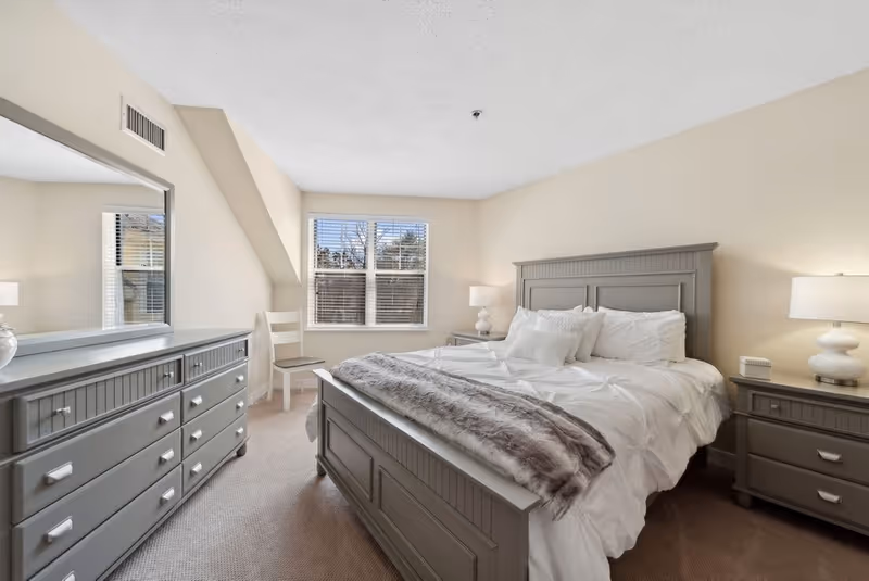 A bright and cozy bedroom featuring a large gray wooden bed with white bedding and a gray fur throw blanket. The room includes matching gray nightstands with white lamps on each side of the bed, a gray dresser with a large mirror, a white chair near the window, and beige walls and carpet. Natural light enters through a window with white blinds.
