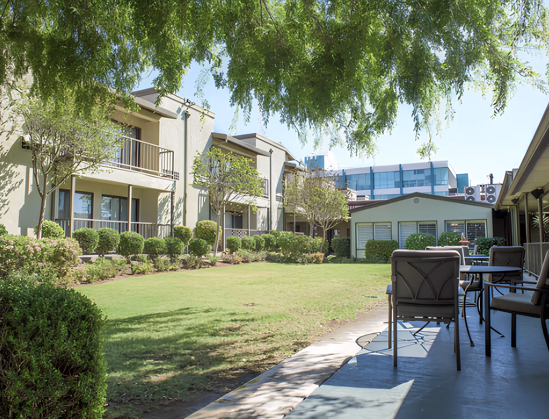 Sunlit courtyard with a lawn, patio seating, and a two-story senior living building surrounded by trees and shrubs.