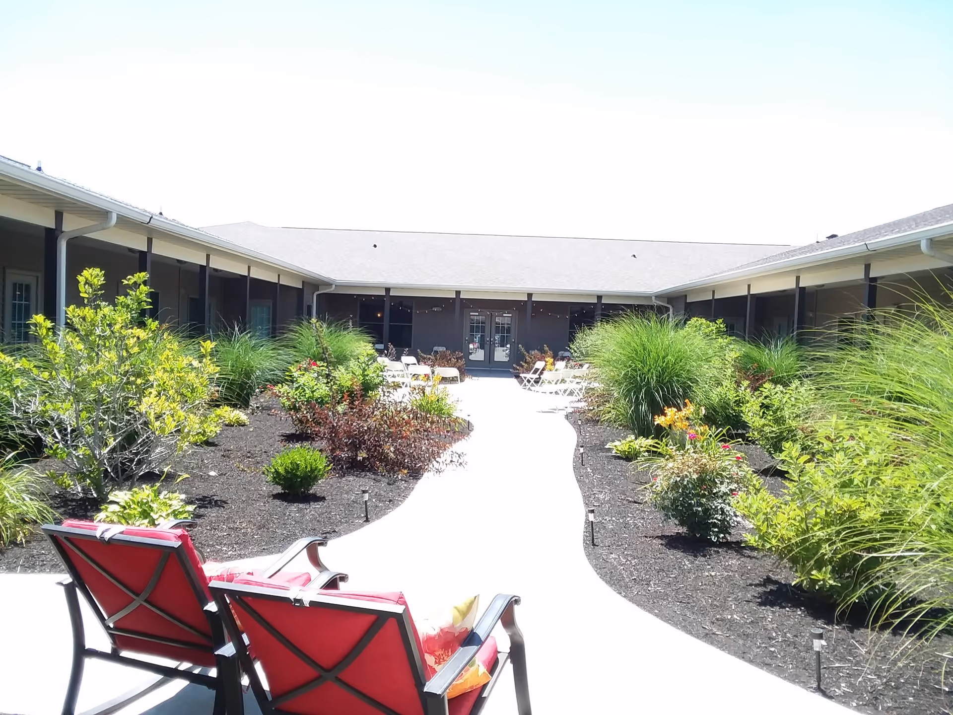 Outdoor courtyard area at Carriage Crossing Senior Living of Champaign featuring a paved walkway lined with various green plants and shrubs. Two red cushioned chairs are positioned in the foreground facing the walkway. The courtyard is surrounded by a single-story building with a covered walkway and glass doors at the far end.