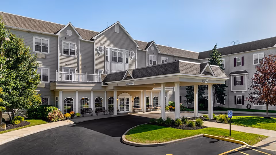Exterior view of Elderwood Assisted Living at West Seneca showing a large, three-story building with gray siding and white trim. The entrance features a covered drop-off area supported by white columns, with benches along the front windows. There are landscaped areas with green grass, trees, and shrubs surrounding the driveway and entrance.