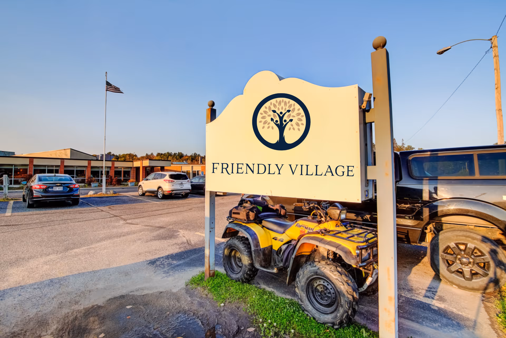A large 'Friendly Village' entrance sign with a tree logo in front of the facility parking lot, vehicles, and an American flag.