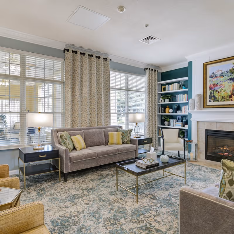 A cozy senior living community lounge with a gray sofa adorned with patterned pillows, two side tables with lamps, a coffee table with books and decorative items, a fireplace with a painting above it, built-in shelves filled with books and decor, and large windows with floral curtains letting in natural light.