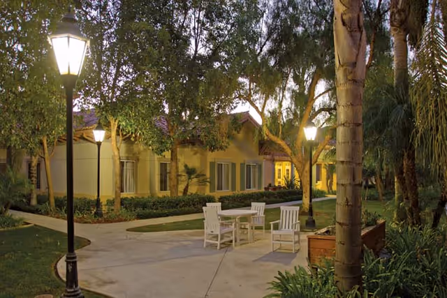 Outdoor patio area at dusk with a round table and four white chairs surrounded by trees, plants, and lit street lamps in front of a single-story building with windows.