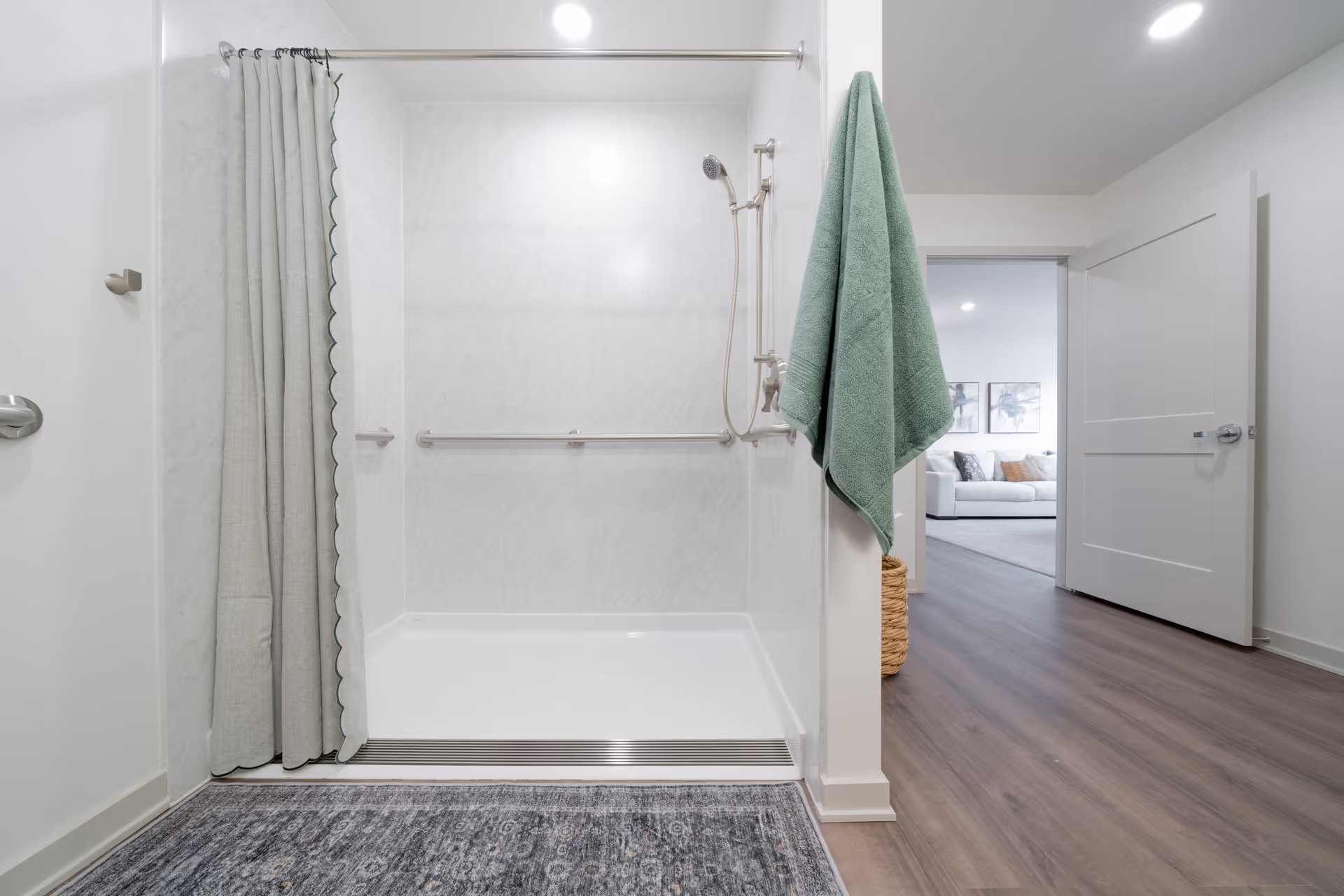 A clean, modern bathroom with a walk-in shower featuring a handheld showerhead and grab bars. A green towel hangs on a hook outside the shower. The bathroom floor has a patterned rug, and the door is open, revealing a living room with a white couch and wall art in the background.