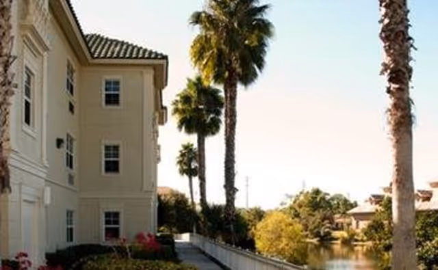 Exterior view of a multi-story beige building with a tiled roof, surrounded by palm trees and landscaping, next to a body of water under a clear sky.