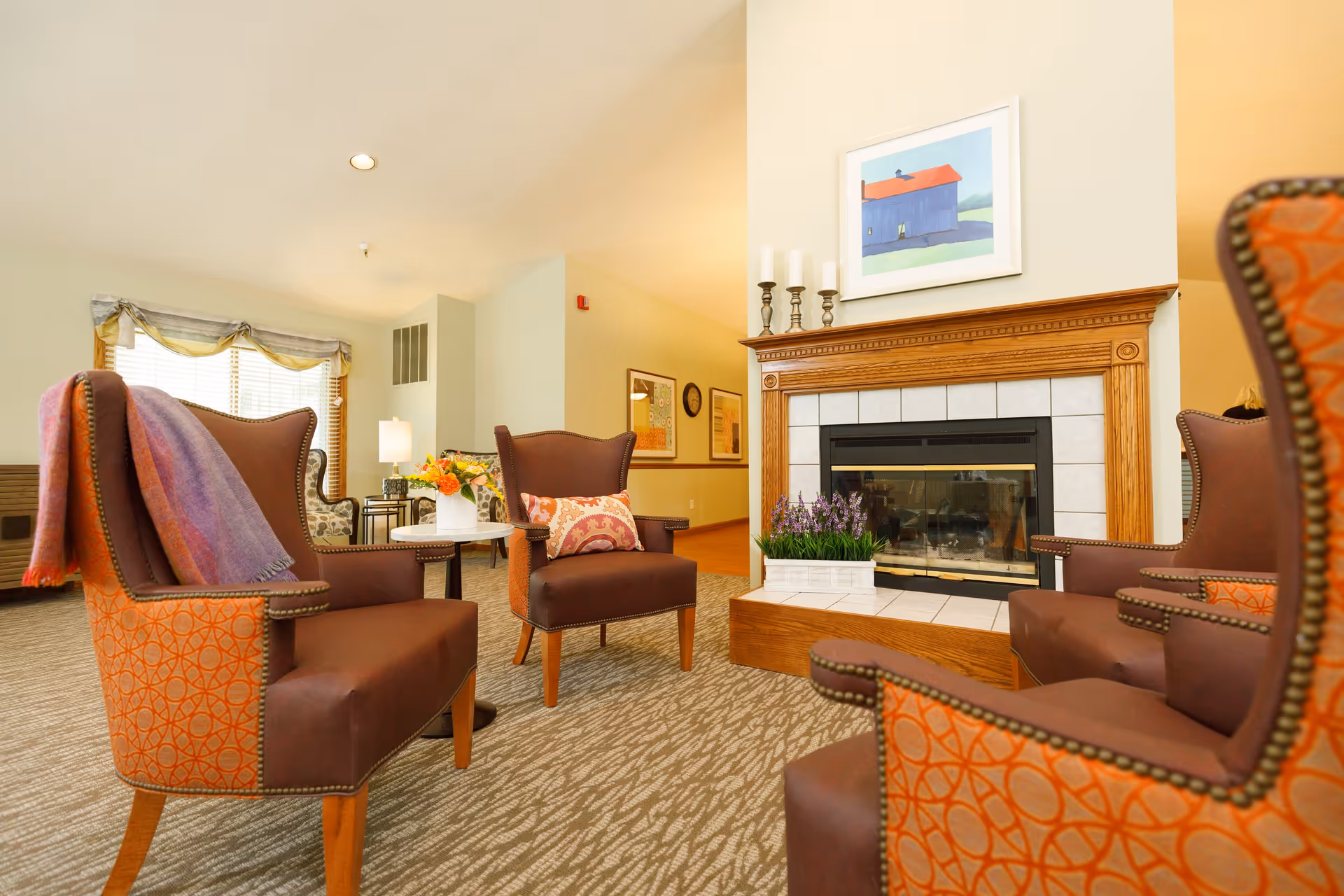A cozy living room area with four brown and orange patterned armchairs arranged around a fireplace. The fireplace has a wooden mantel with three white candles and a framed painting of a blue barn with a red roof above it. A small white table with a vase of flowers is placed between two of the chairs. The room has beige walls, carpeted flooring, and a window with blinds and a valance in the background.
