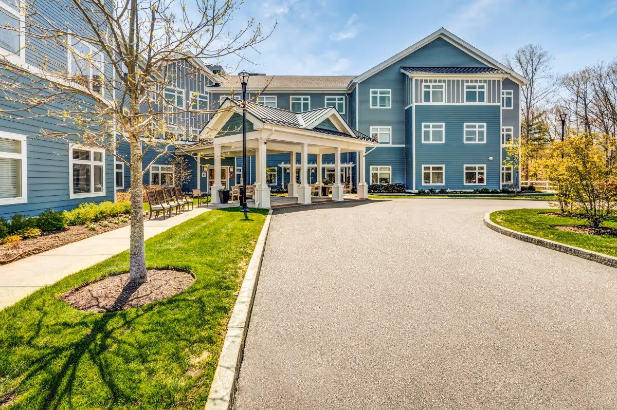 Exterior view of a senior living facility with a blue three-story building, a covered entrance with white pillars, a paved driveway, green lawns, trees, and benches along the sidewalk.