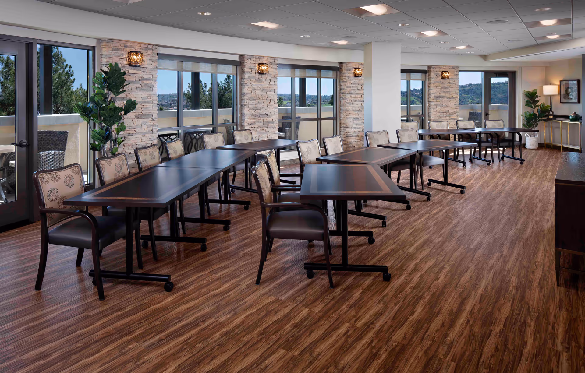 A spacious, well-lit room with multiple tables and chairs arranged in rows on a wood floor. Large windows and glass doors line one side, allowing natural light and views of trees and hills outside. Stone pillars and modern wall sconces add decorative elements, with potted plants and a side table with a lamp in the background.