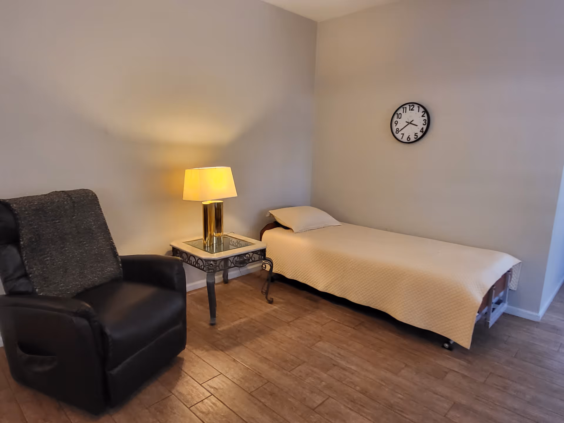 A simple room with a single bed covered with a beige quilt, a pillow, a side table with a lit lamp, and a black armchair with a gray blanket draped over it. A round wall clock is mounted on the wall above the bed.
