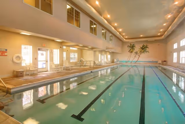Indoor swimming pool with clear water and lane markings, surrounded by a tiled deck with chairs and handrails. The room has high ceilings with recessed lighting and windows along the walls. A mural of palm trees is painted on the far wall.