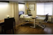 A single hospital-style bed in a well-lit room with wooden flooring. The bed has white sheets and is positioned near a window with blinds. There is a small table on wheels next to the bed, a chair and desk on the left side, and a curtain that can be drawn for privacy. A vase with flowers and a framed picture hang on the wall above the bed.