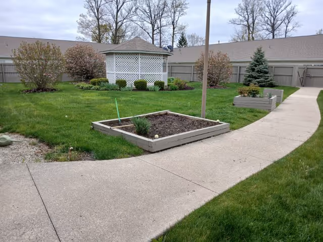 A landscaped outdoor area with a concrete pathway curving through green grass. There is a white gazebo with lattice sides in the background, surrounded by bushes and small trees. Raised garden beds are visible along the pathway, and a wooden fence encloses the area.