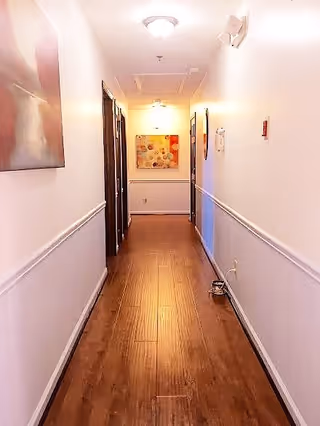 A well-lit hallway with wooden flooring and white walls, decorated with framed artwork on both sides. There are several doorways along the hallway and a ceiling light fixture.