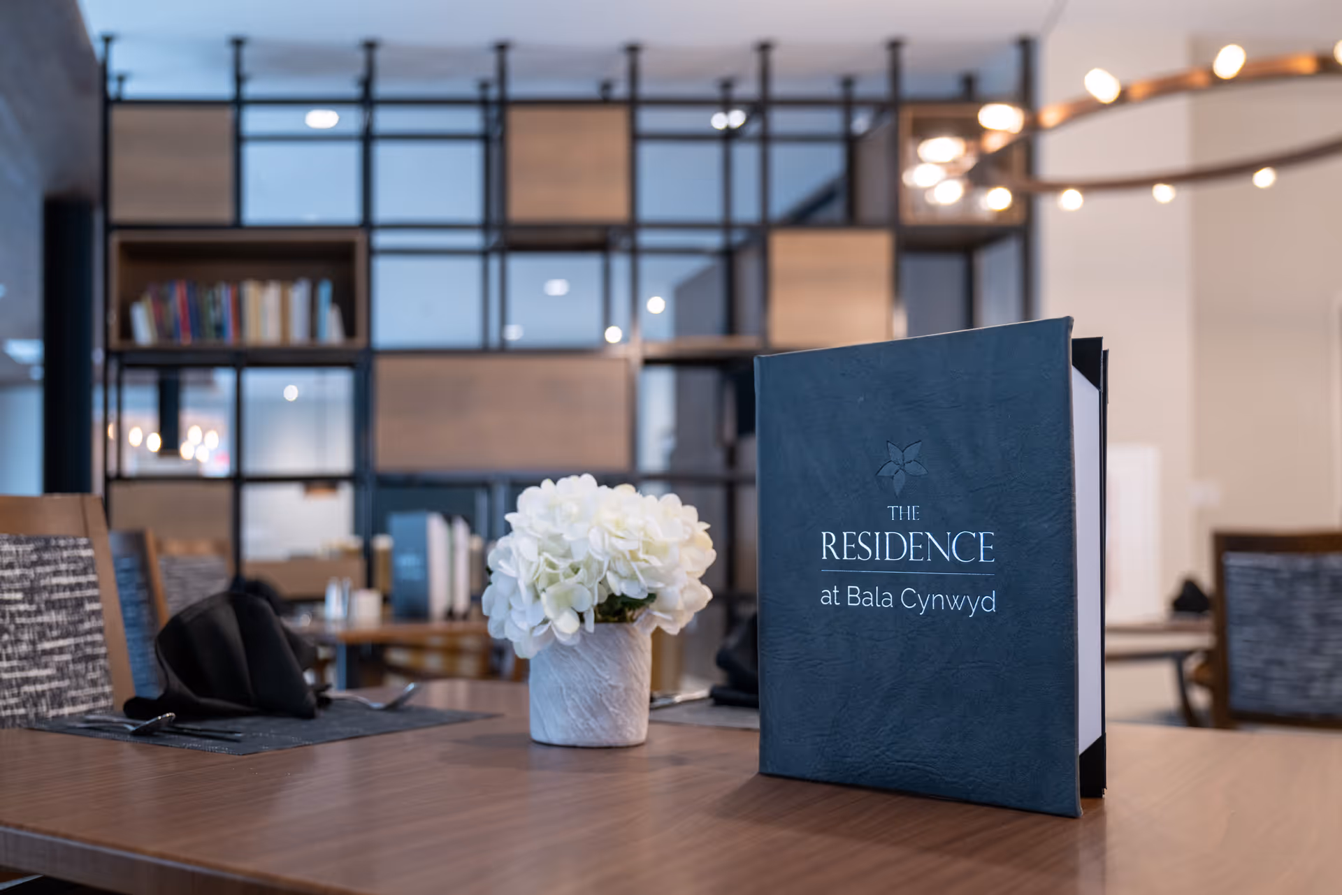 Dining table with a menu reading 'The Residence at Bala Cynwyd' and a small vase of white flowers in a modern dining area.