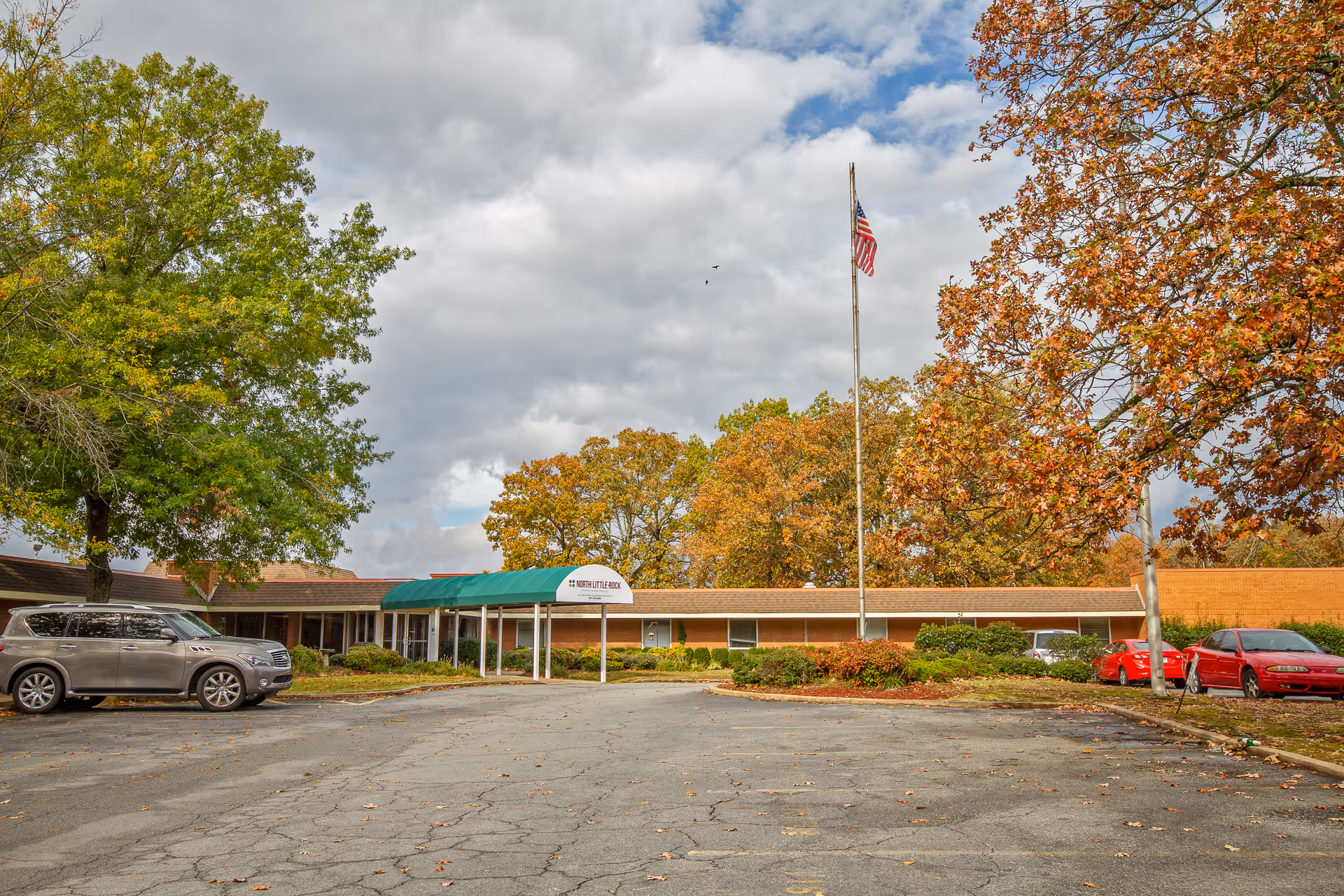 Front exterior of a single-story nursing center with a covered entrance, an American flag on a pole, parked cars, and autumn trees.
