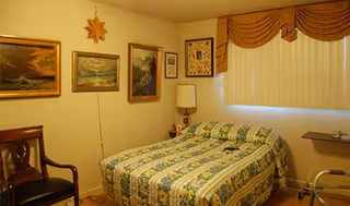 A small bedroom with a bed covered in a green and white patterned bedspread. There are three framed paintings on the wall above the bed and a lamp on a small bedside table. A wooden chair is placed against the opposite wall, and a window with vertical blinds and a brown valance is visible. A walker is positioned near the bed.