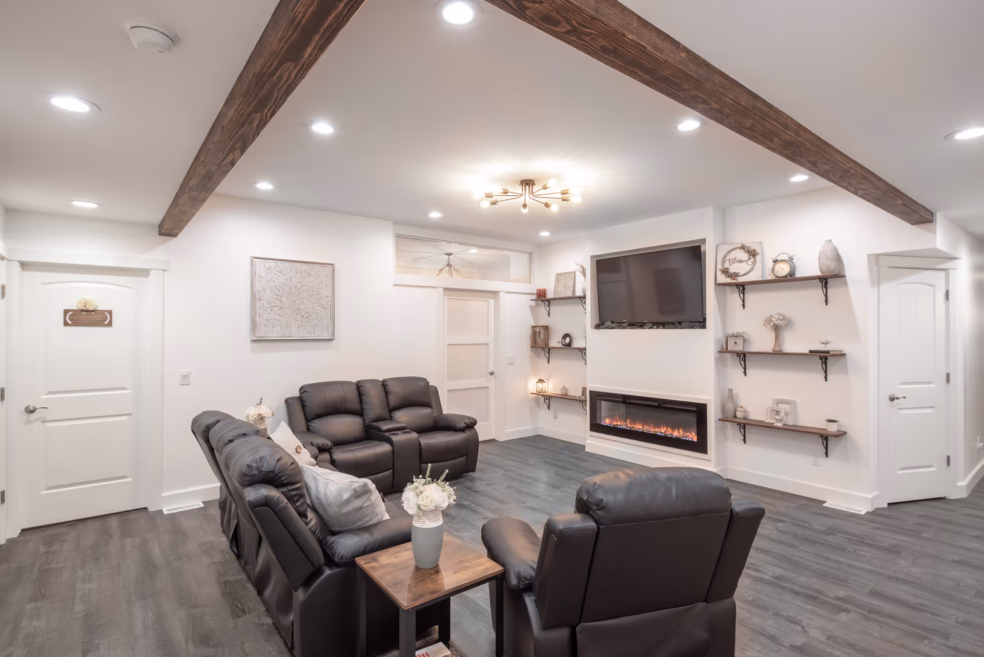 A cozy senior living room with dark leather recliner chairs arranged around a wooden side table with a flower vase. The room features a modern electric fireplace beneath a mounted flat-screen TV, white walls with decorative shelves holding various ornaments, and exposed wooden ceiling beams. The floor is dark wood, and there are two white doors on either side of the room.