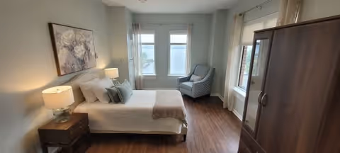 Sunlit bedroom with a made bed, bedside tables and lamps, a patterned armchair by two windows, and a wooden wardrobe.