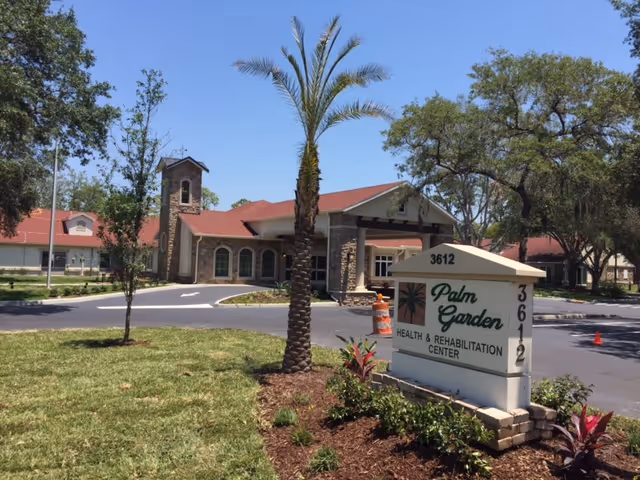 Exterior view of Palm Garden Health & Rehabilitation Center with a clear blue sky, a driveway, palm trees, and a sign displaying the facility's name and address.