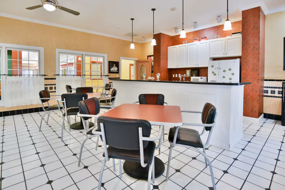 Communal dining area with red-topped tables and black chairs facing a white kitchenette/bar.