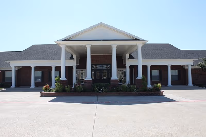 Front exterior view of a single-story brick building with white columns supporting a covered entrance, surrounded by a paved driveway and some landscaping with shrubs and flowers.