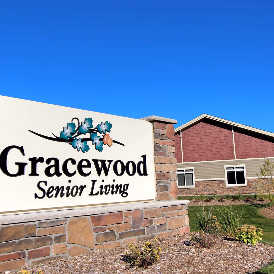 Outdoor view of the Gracewood Senior Living facility sign with a stone base and decorative leaves above the text. In the background, there is a building with beige and red siding under a clear blue sky.