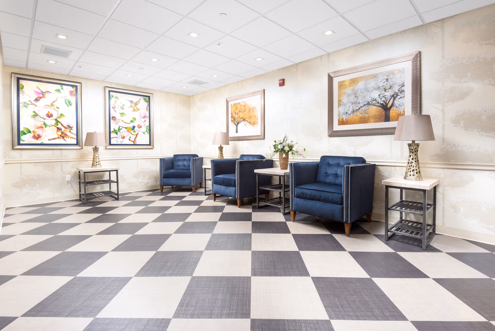 A bright and clean sitting area with four navy blue armchairs arranged along the wall. Each chair is paired with a small side table topped with a decorative lamp. The floor features a large checkered pattern in shades of gray and white. The walls are adorned with framed artwork depicting colorful birds and trees with autumn foliage.