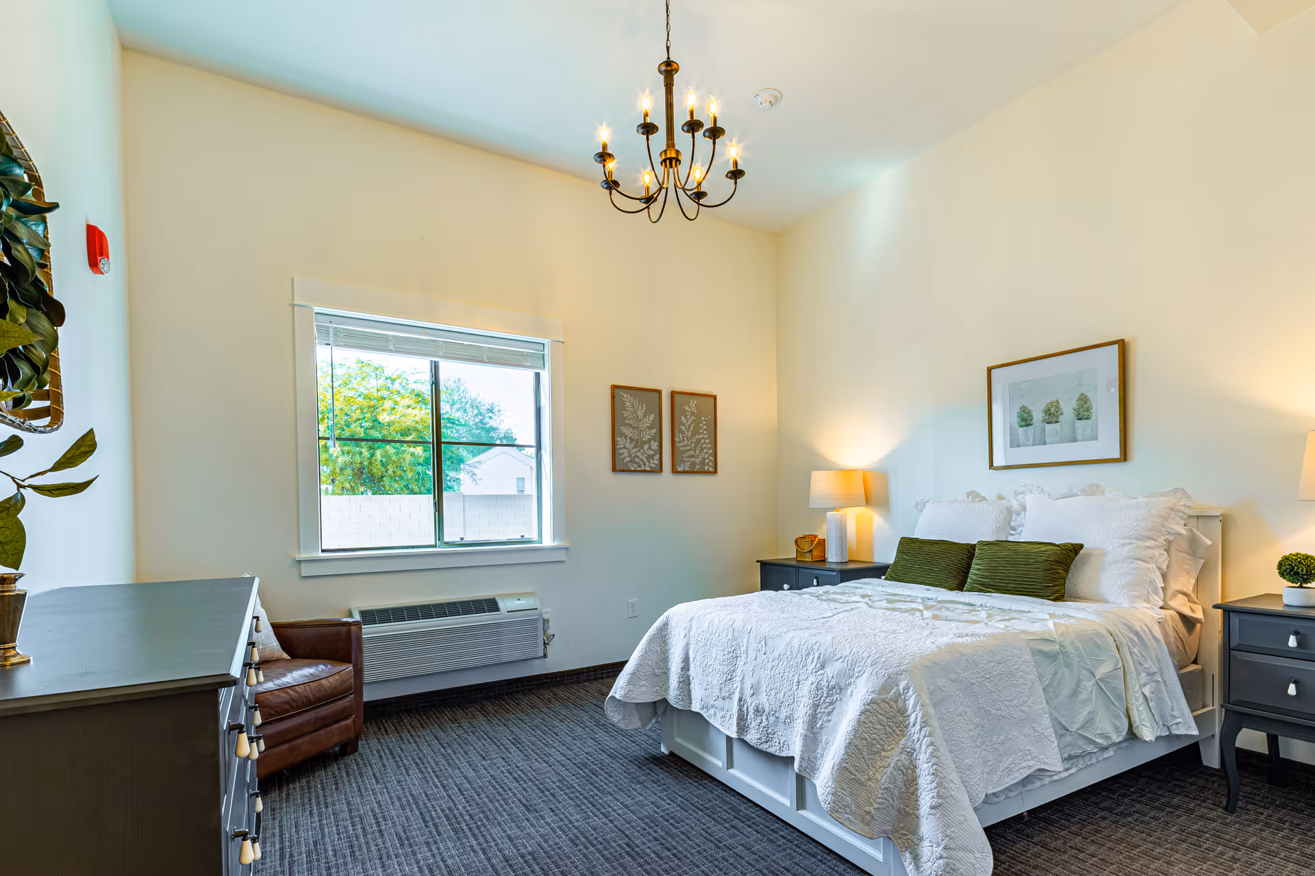 A bright furnished bedroom with a bed, nightstands, chandelier, dresser, window, and armchair.