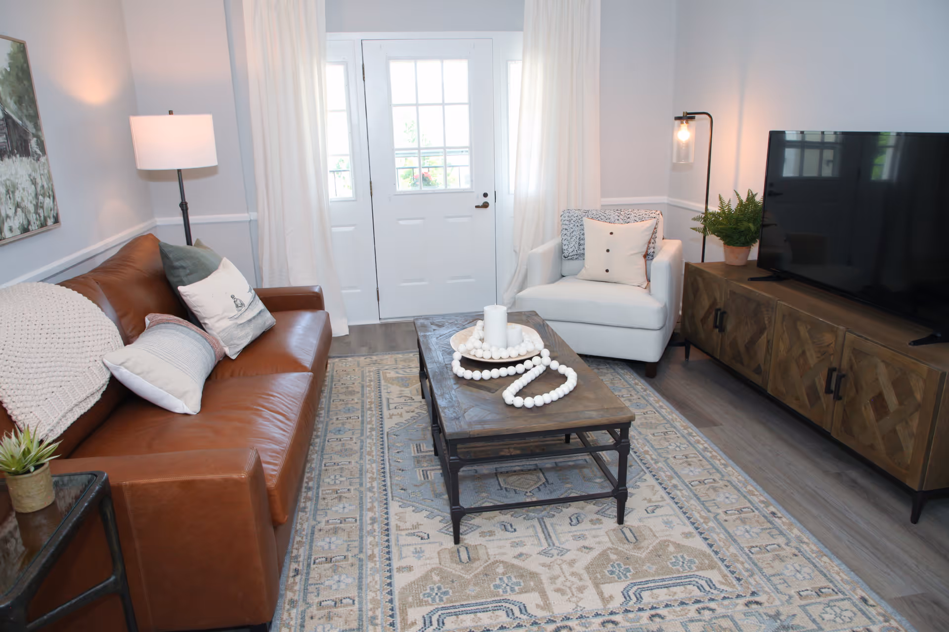 A cozy living room with a brown leather sofa adorned with pillows and a knitted throw, a white armchair with cushions, a wooden coffee table with decorative beads and a candle, a patterned area rug, a wooden TV stand with a flat-screen TV, a potted plant, a floor lamp, and a door with windows letting in natural light.