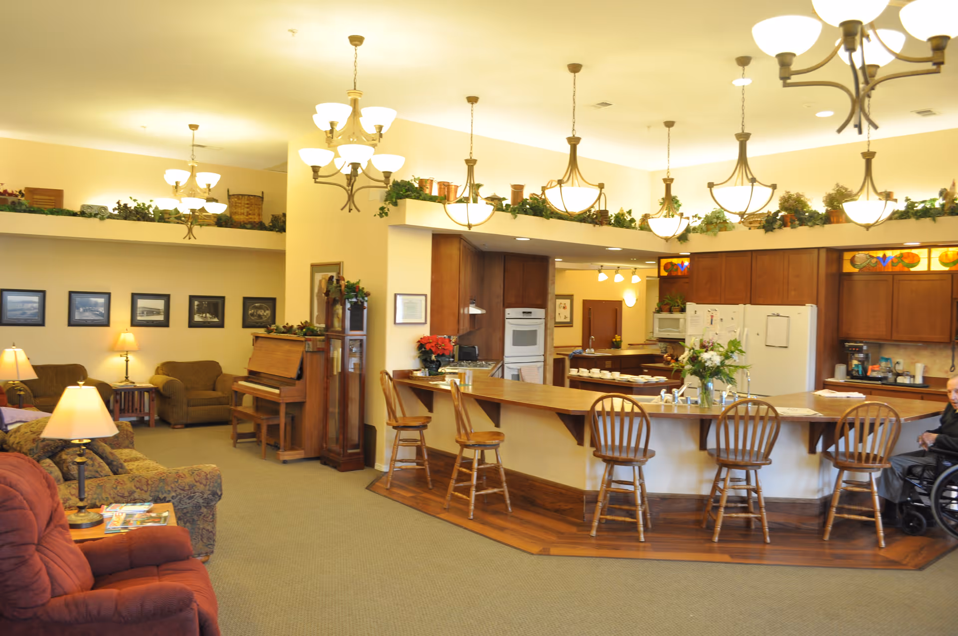 A spacious common area in a senior living facility featuring a kitchen with wooden cabinets, a large counter with bar stools, and a living room area with comfortable armchairs and a piano. The room is warmly lit with multiple hanging light fixtures and decorated with plants and framed pictures on the walls. A person in a wheelchair is visible near the kitchen counter.