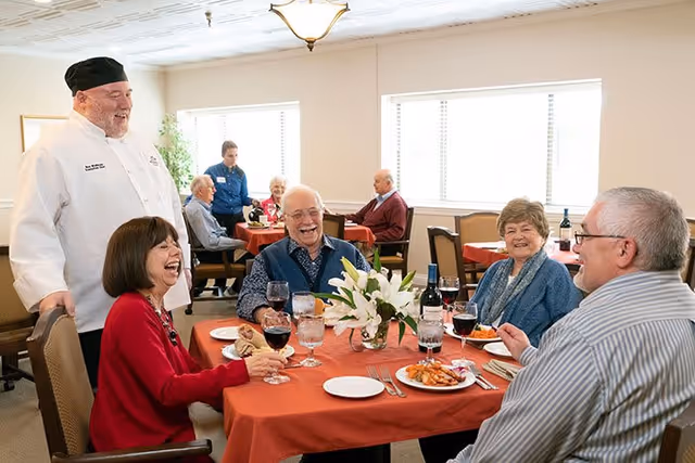 A group of elderly people sitting around a dining table with red tablecloths, enjoying a meal and drinks. A chef in white uniform and black hat stands beside the table smiling. The room is well-lit with large windows and other diners are visible in the background.