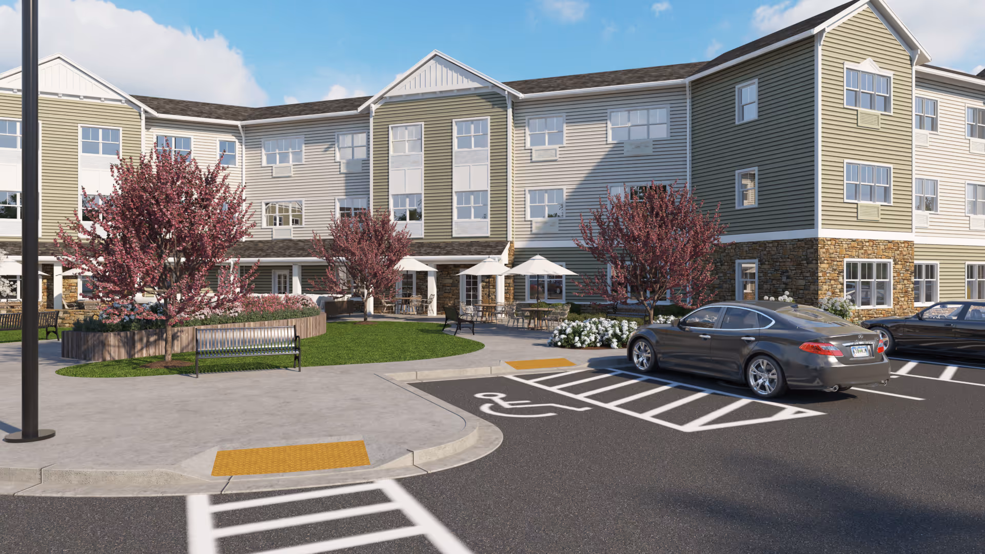 Front exterior of a three-story senior living building with a landscaped courtyard, benches, patio seating, and parked cars.