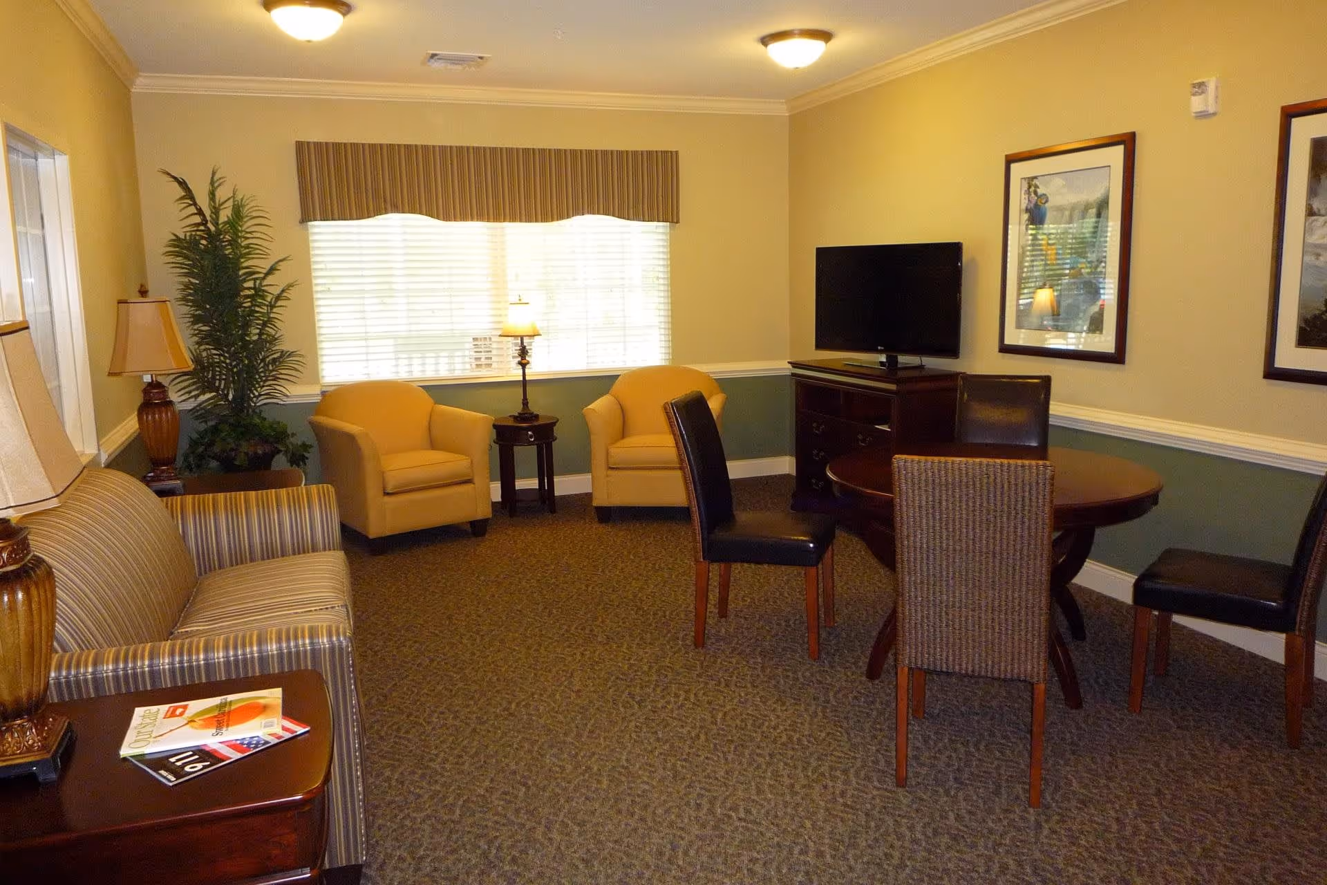 A cozy living room with a striped sofa, two yellow armchairs, a round wooden table with four chairs, a flat-screen TV on a wooden cabinet, two framed pictures on the wall, a potted plant, and two table lamps. The room has beige walls with a green lower half and a window with blinds and a striped valance.