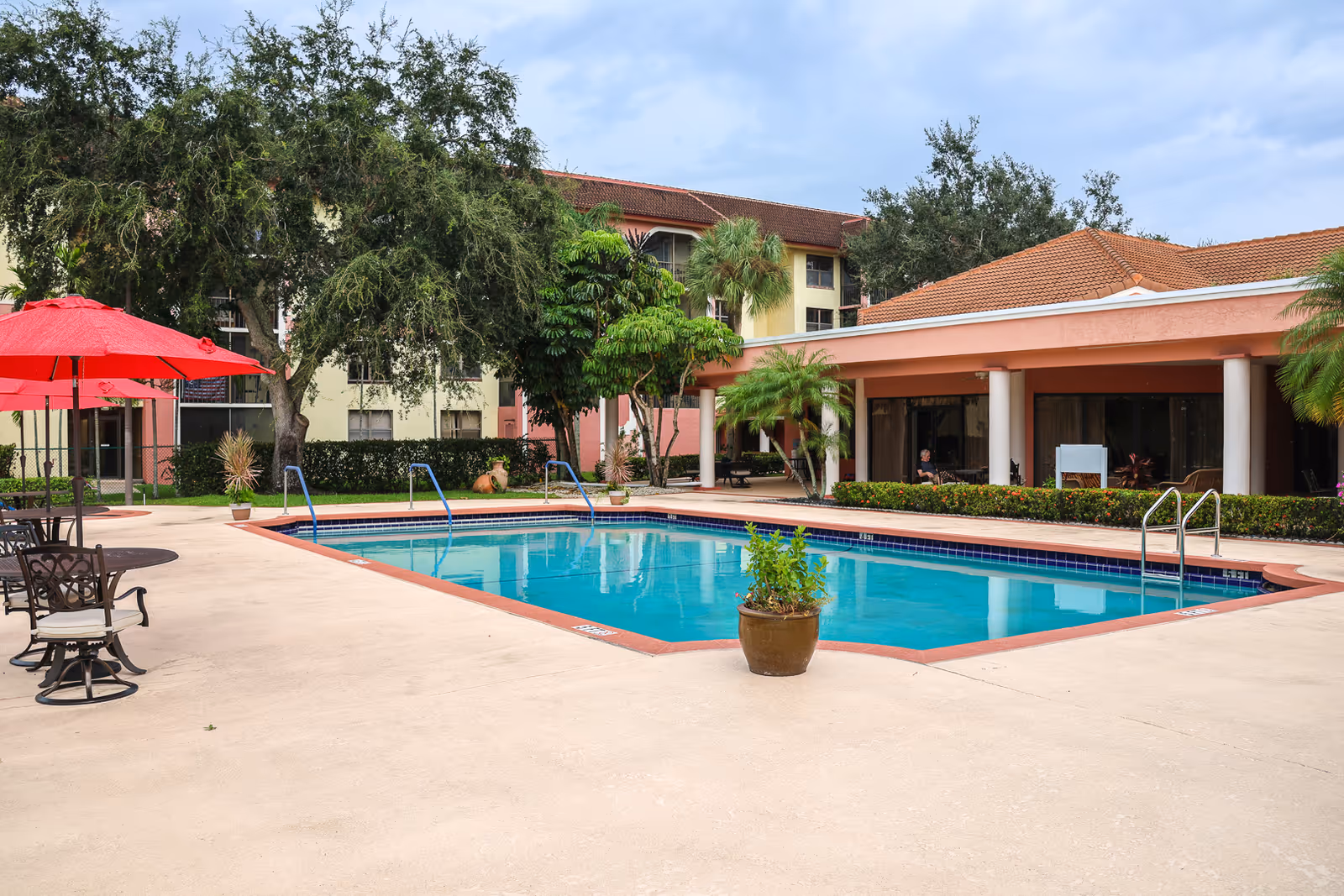Outdoor swimming pool area at Elison Independent Living of Lake Worth with a clear blue pool, surrounded by a concrete deck. There are red umbrellas and metal chairs on the left side, lush green trees and plants around the pool, and a building with a tiled roof in the background.