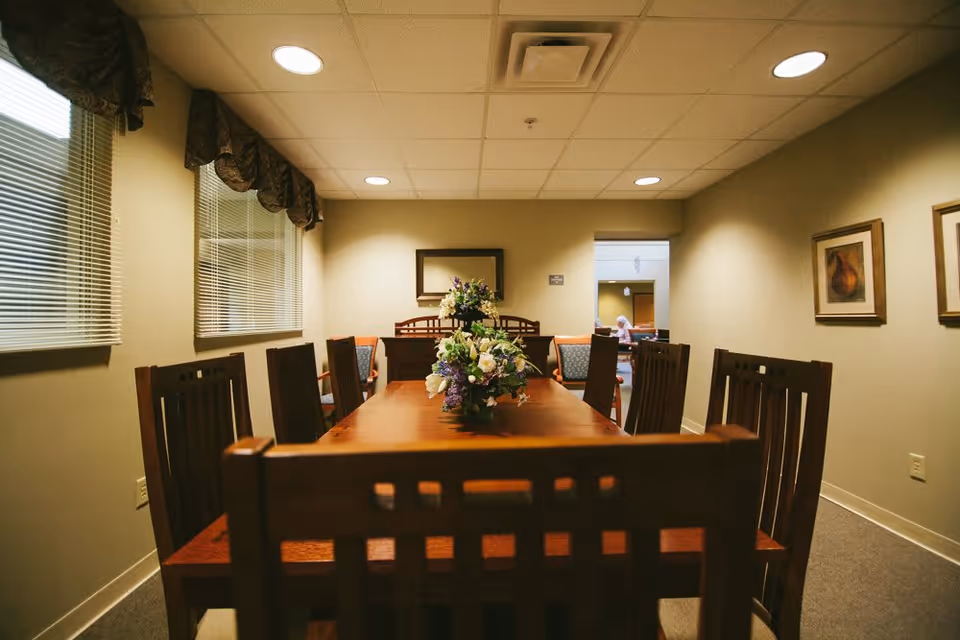 A dining room with a long wooden table surrounded by wooden chairs. There are two floral centerpieces on the table. The room has beige walls, two windows with blinds and valances on the left, and framed artwork on the right wall. Recessed ceiling lights illuminate the room, and an open doorway at the back reveals another room with a person sitting.